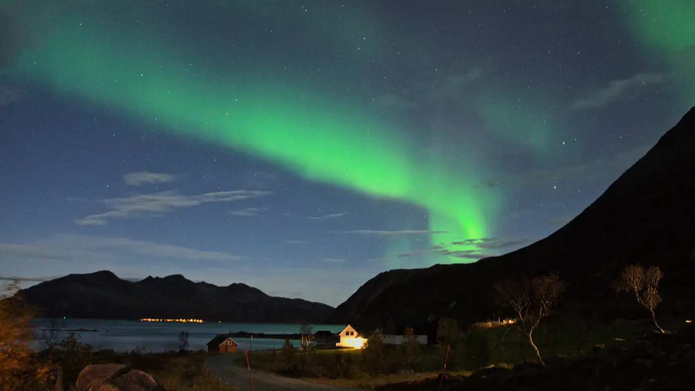 A vivid green aurora borealis sweeps across the night sky above a Norwegian fjord village with illuminated cabins and mountains silhouetted in the foreground
